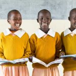 Three African girls in bright yellow sweaters laughing as they stand in front of the class presenting from their books.